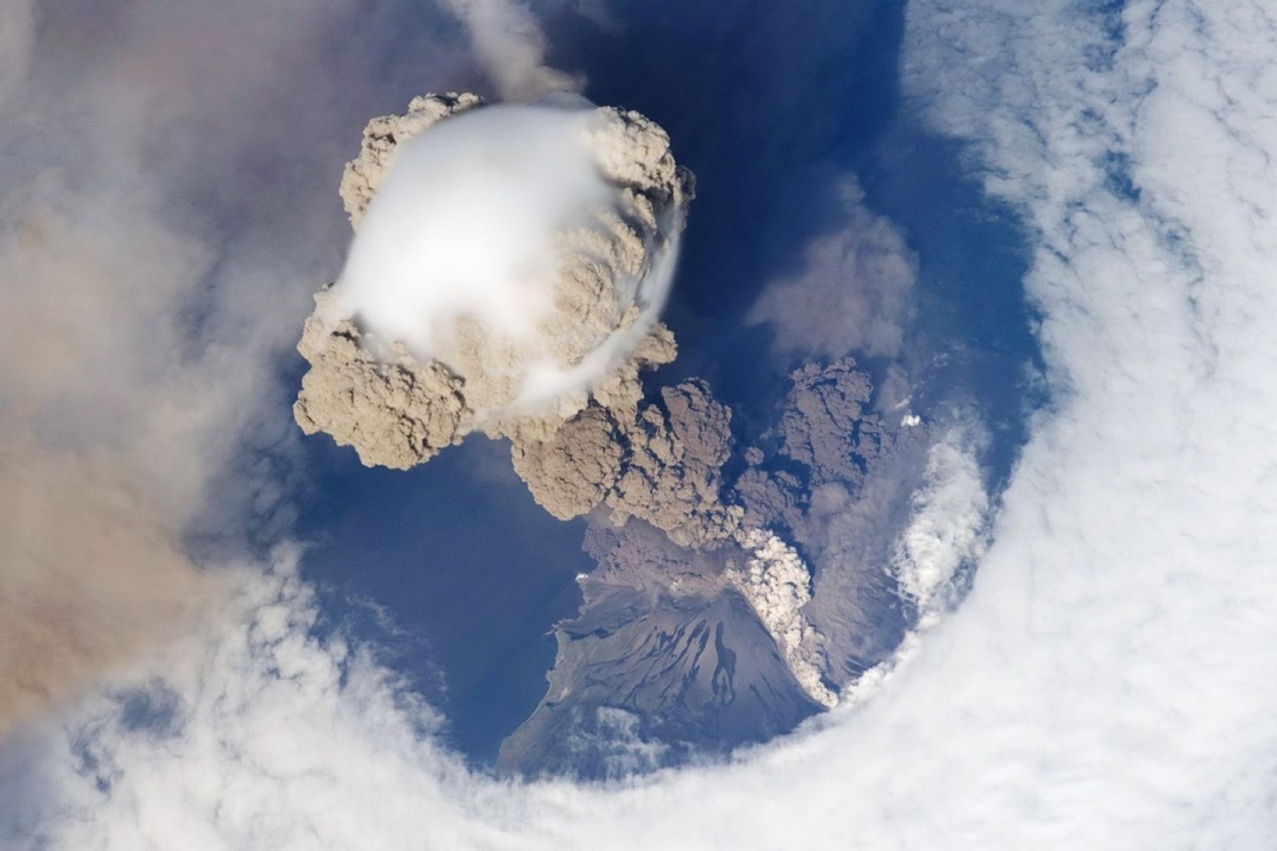 Astronaut photograph of an eruption plume rising through clouds above Sarychev Peak in the Kuril Islands.